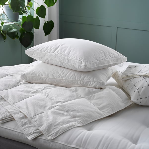 Close-up of a white mattress with a pile of white duvets and pillows on top of it. Green wall and window with green plant.