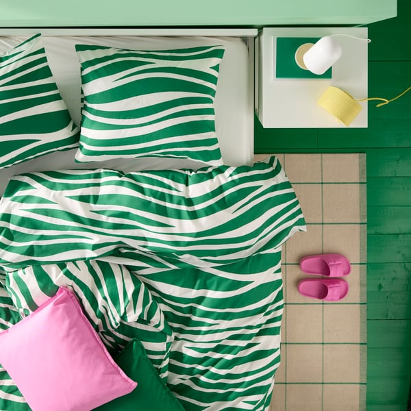 A desk lamp and a Bluetooth speaker sit on a bedside table placed next to a bed with green and white PORTLAKMÅLLA bed linen.