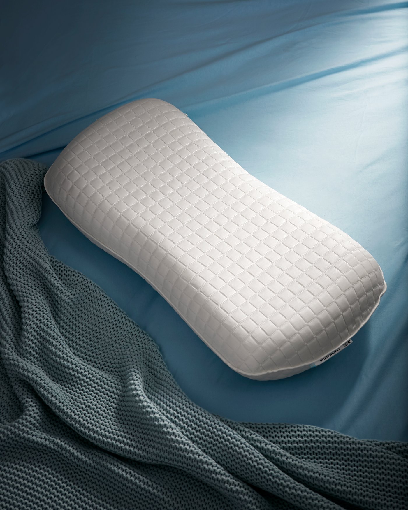 Almohada ergonómica IKEA sobre una cama con sábanas celestes y plumón gris.