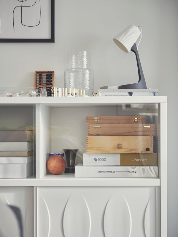 Puzzles and games behind the glass of a white KALKNÄS cabinet with sliding doors. On top is a SVALLET lamp and more games.