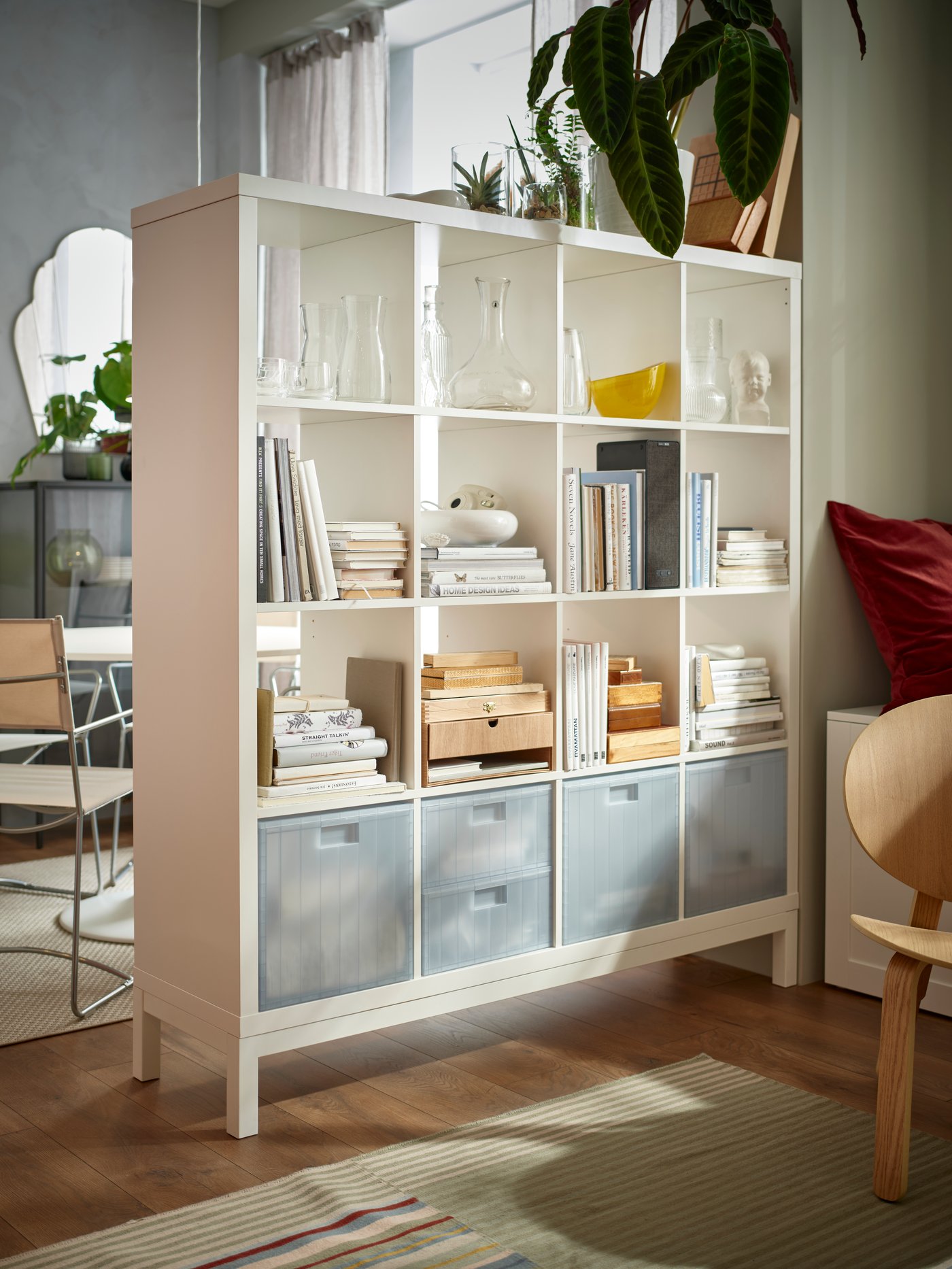 Une étagère blanche KALLAX à compartiments remplis de livres, objets déco et boîtes de rangement se trouve dans une pièce lumineuse avec une fenêtre, des plantes au-dessus et des meubles autour.

