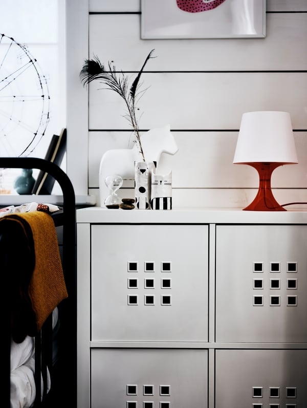 Une étagère KALLAX avec une lampe rouge, un cheval blanc et un vase posés dessus, à côté d’un cadre de lit devant un mur blanc.