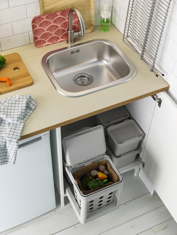 A door under the sink is open, revealing light grey waste sorting baskets that slide out and small stacked baskets.