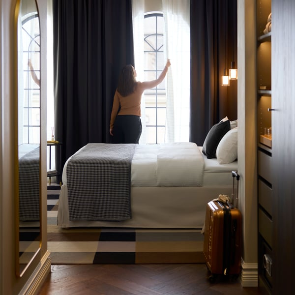 A person stands by dark gray KORGMOTT blackout curtains that partially cover thin curtains at a large, sunny hotel room window.