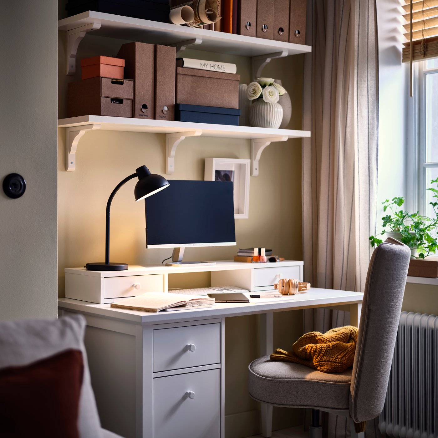 A corner with a study space centred around a white GULLABERG desk with a drop-file drawer, softly lit by a desk lamp.