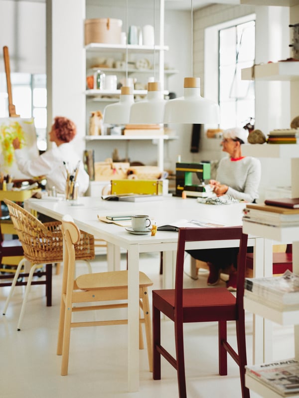 Two people in a dining area with assorted chairs around four white MELLTORP tables below three BUNKEFLO pendant lamps.