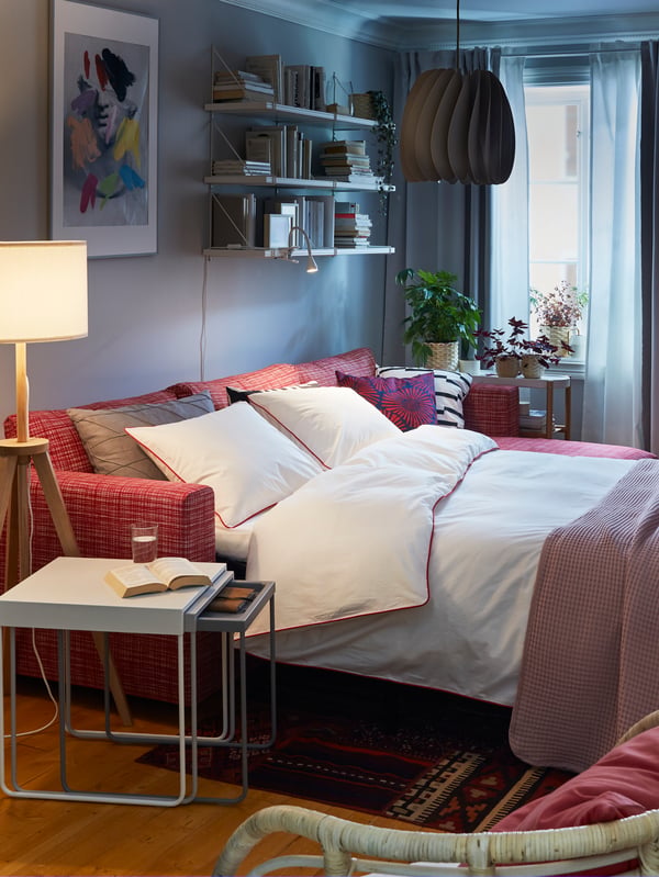 Living room interior with sofa-bed in double-bed mode. Around it: a nest of tables, seating, wall-mounted bookcase with books.
