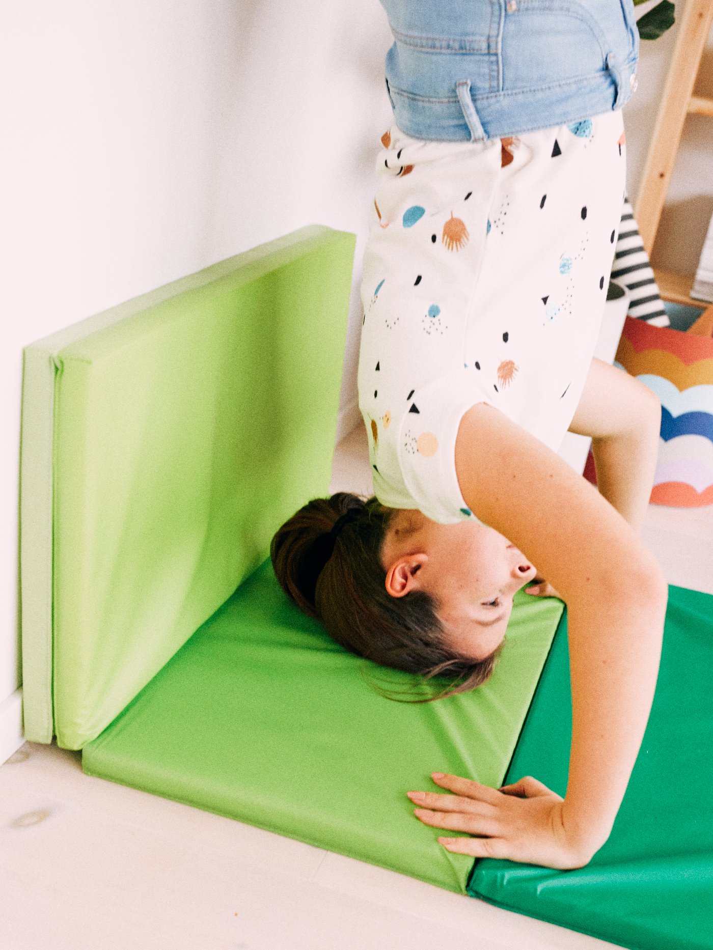 A young girl performs a handstand against a wall, her head and arms supported by a bright green PLUFSIG folding gym mat.