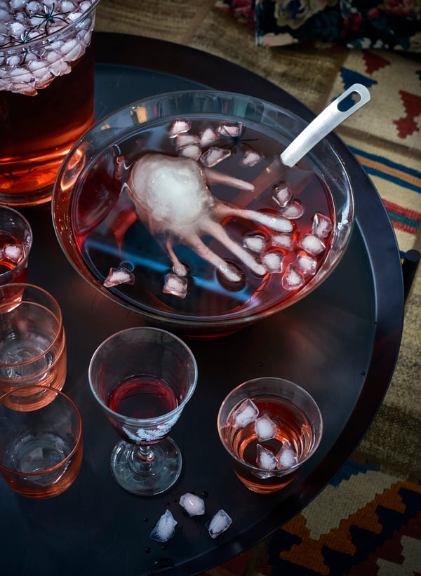 A punch bowl with red punch filled with ice cubes and one human hand shaped ice cube, sitting on a black table with glasses.
