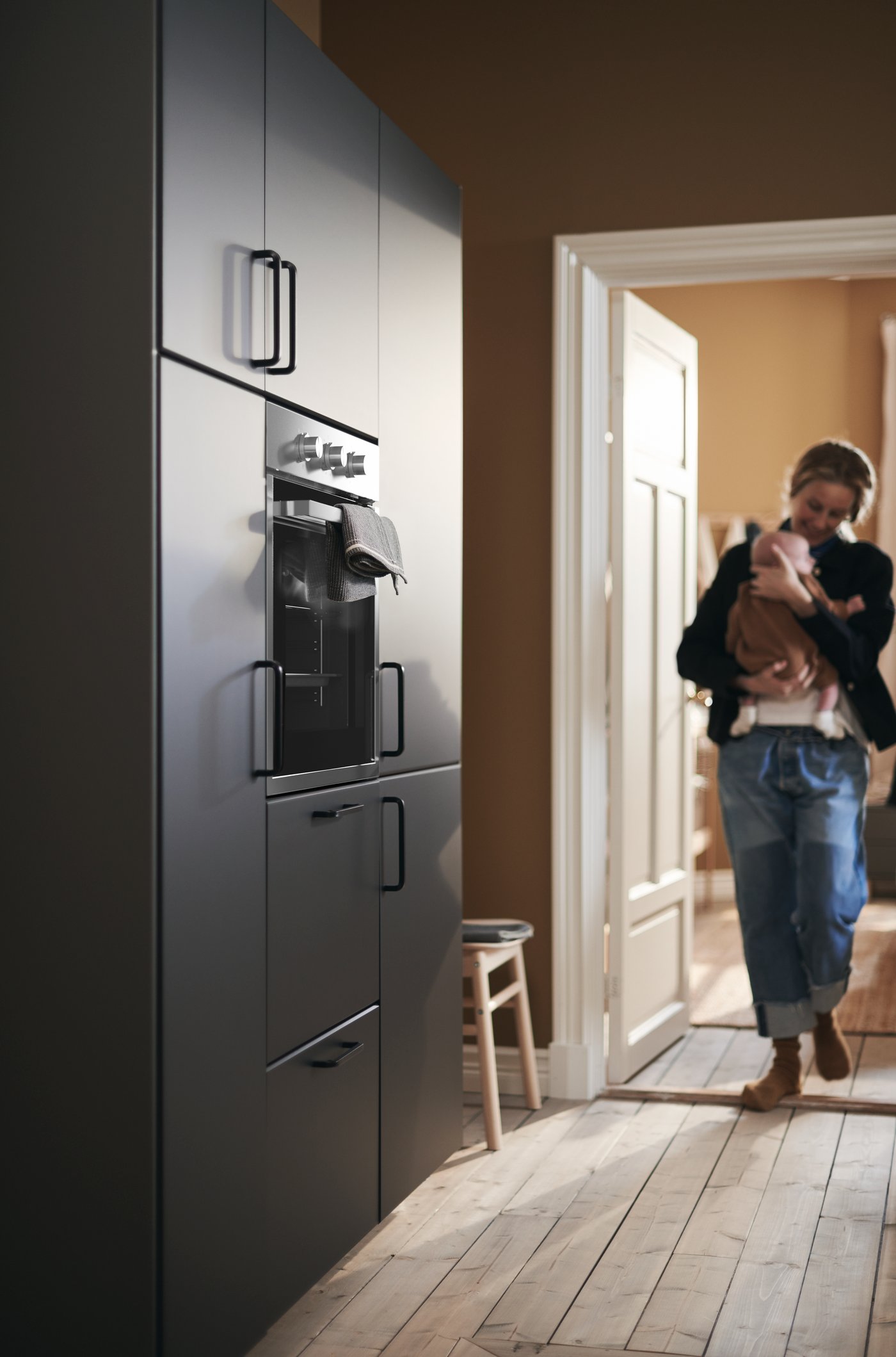 A mother cradles her baby as she enters the kitchen, approaching a wall of cabinets with KUNGSBACKA fronts.