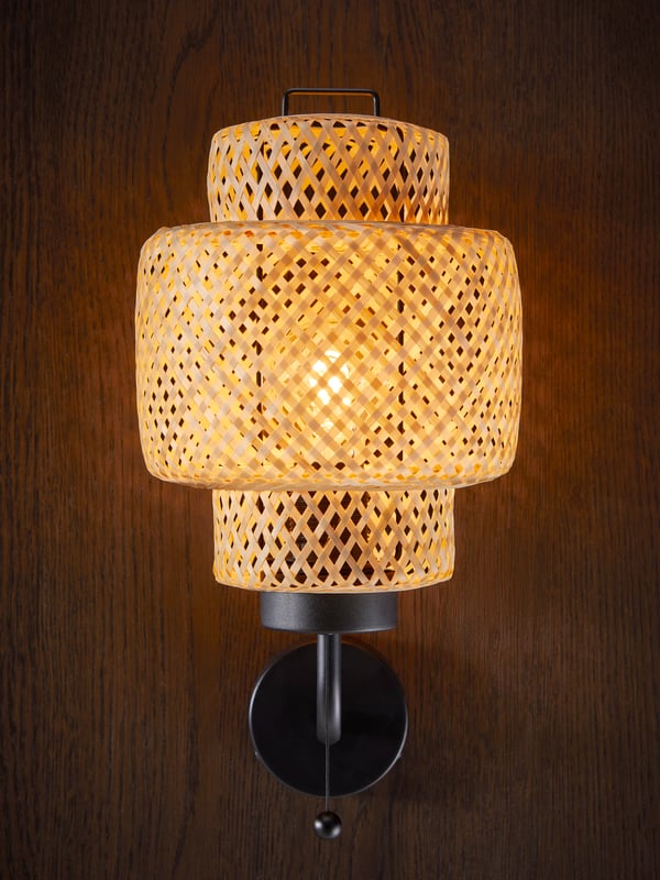A lit braided-bamboo SINNERLIG wall lamp with wired-in installation is mounted on a wall made of dark wood.