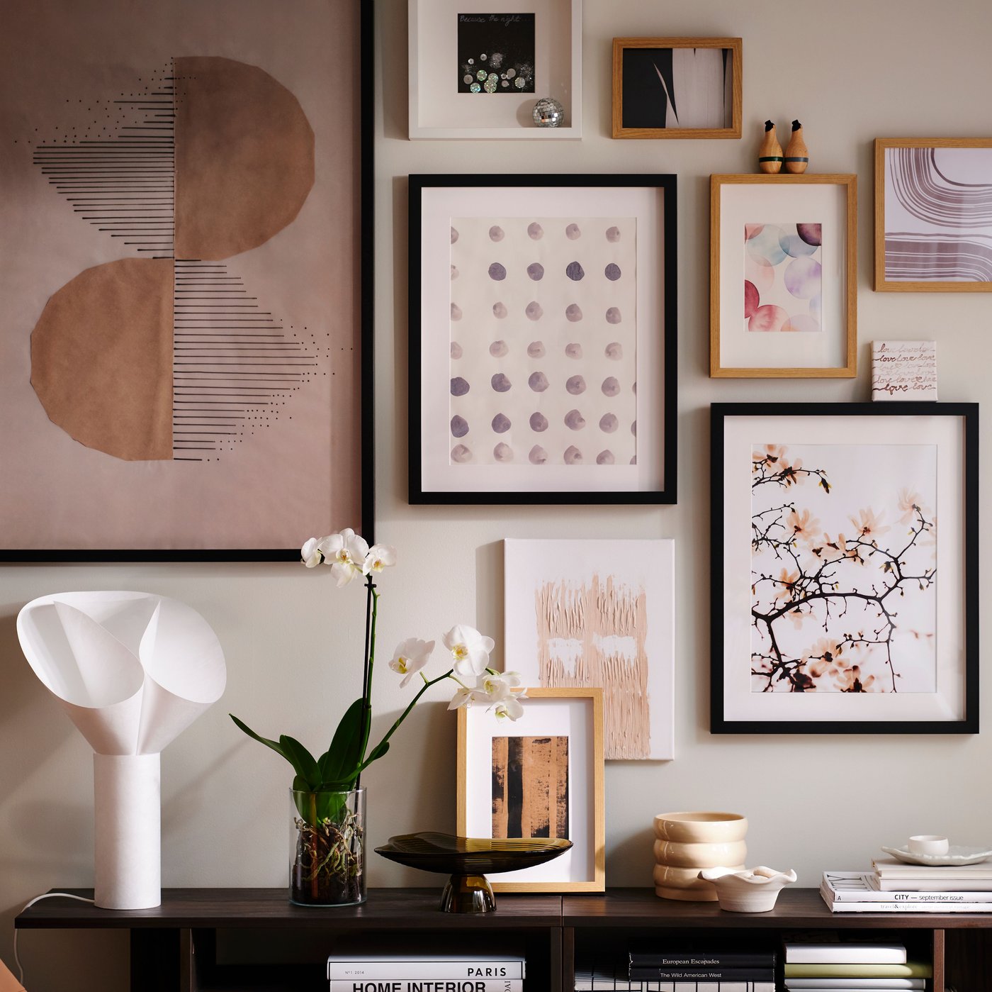 A wall in a light living room with an art cluster in various RÖDALM frames above HOLMERUD side tables with orchids and deco.