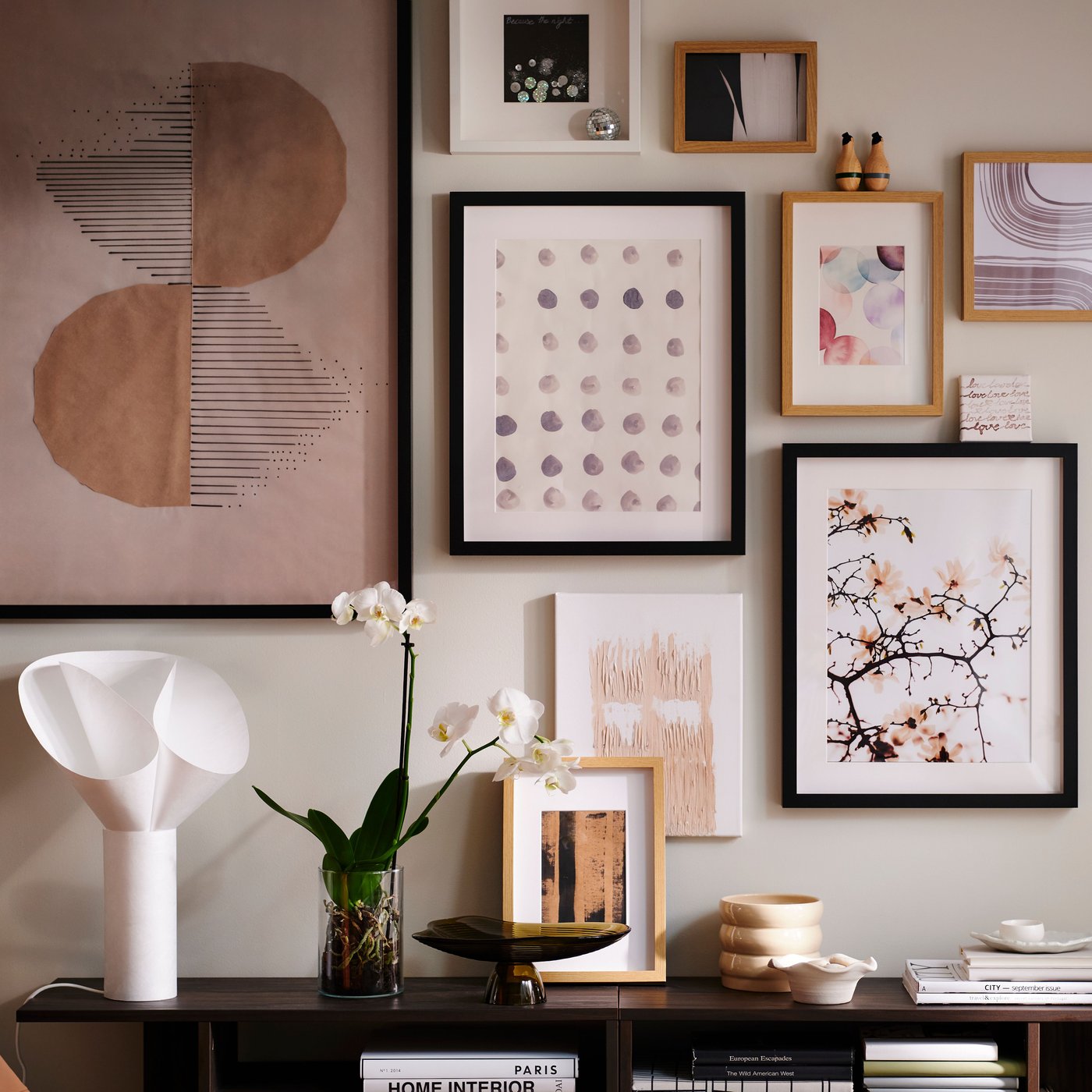 A wall in a light living room with an art cluster in various RÖDALM frames above HOLMERUD side tables with orchids and deco.