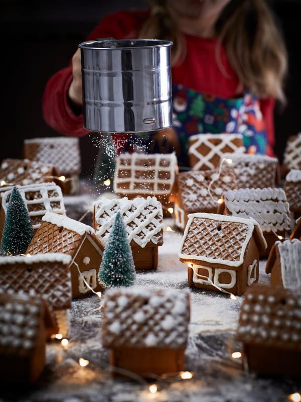Una colección de casitas de jengibre en miniatura dispuestas en forma de pueblecito con árboles verdes de cepillo de botella y luces centelleantes entre las casas.