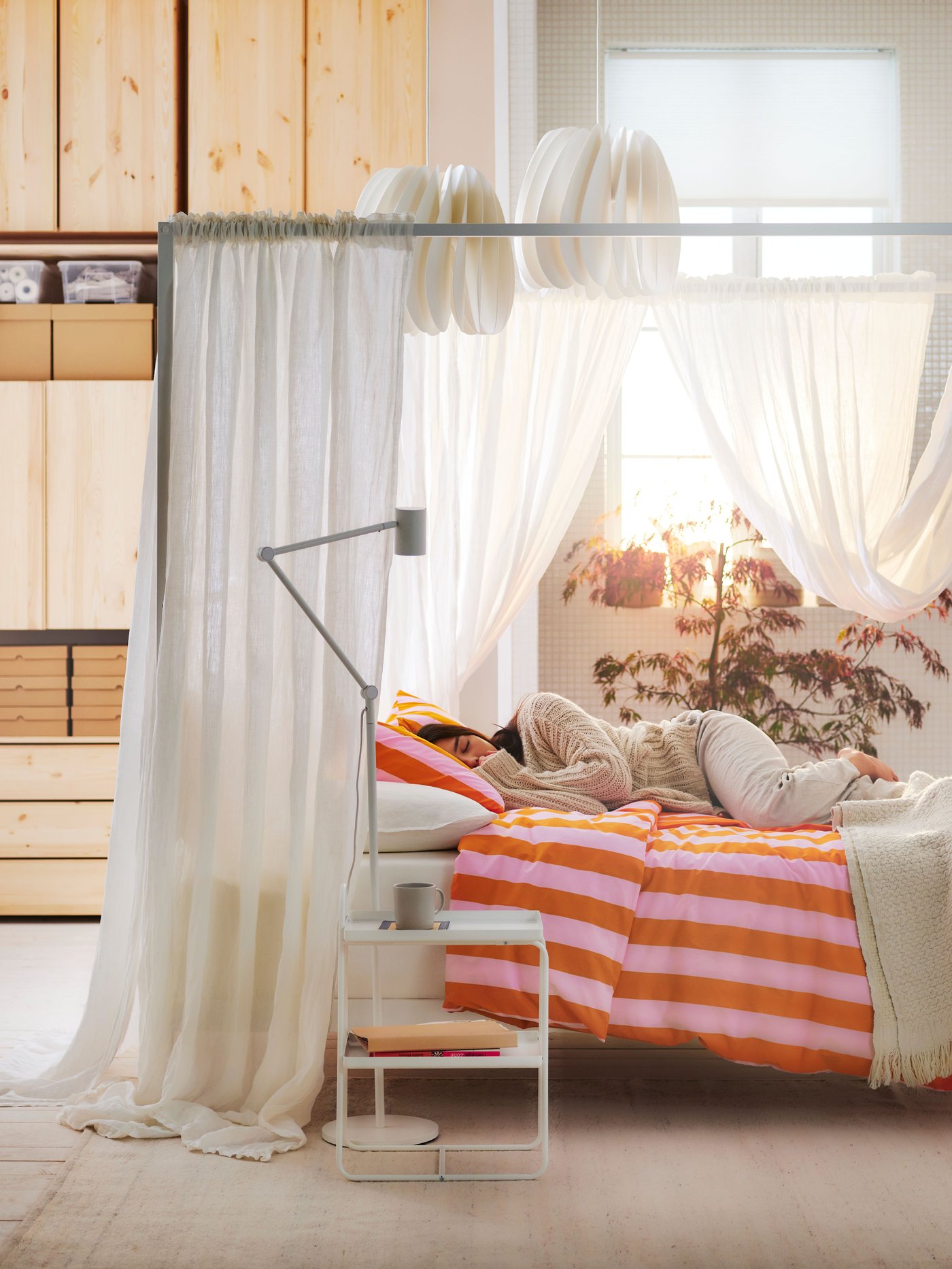 Ett sovrum med en himmelssäng draperad i vita skira gardiner, där en person ligger och vilar på en säng bäddad med orange‑ och rosalrandigt sänglinne. En vit sänglampa och ett sidobord står intill.
