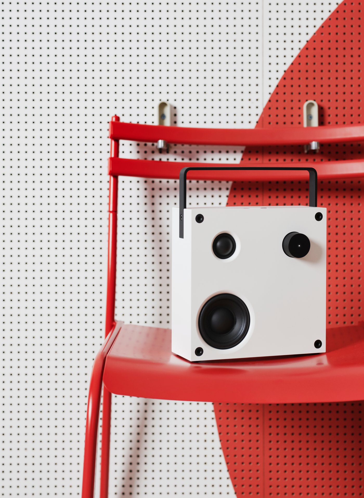 A white VAPPEBY speaker on top of a red GENESÖN metal chair that is hanging on two hangers mounted on a white and red wall.