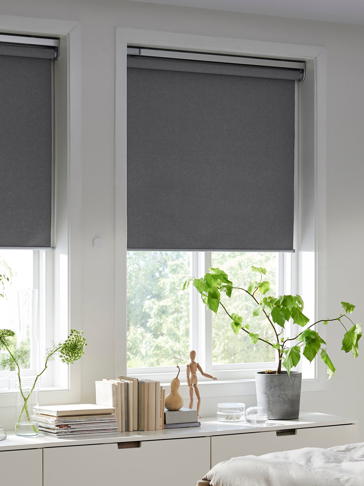 Dos ventanas largas con cortinas roller grises a medio bajar. Debajo de las ventanas hay una cómoda blanca con cajones.