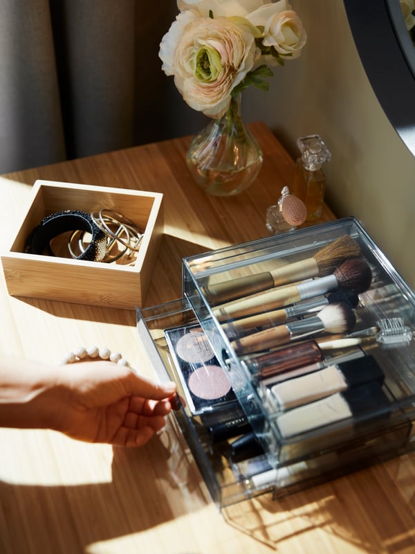 A smoked GODMORGON mini chest with 2 drawers with makeup items, a TAVELÅN tray with jewellery on a bamboo dressing table.