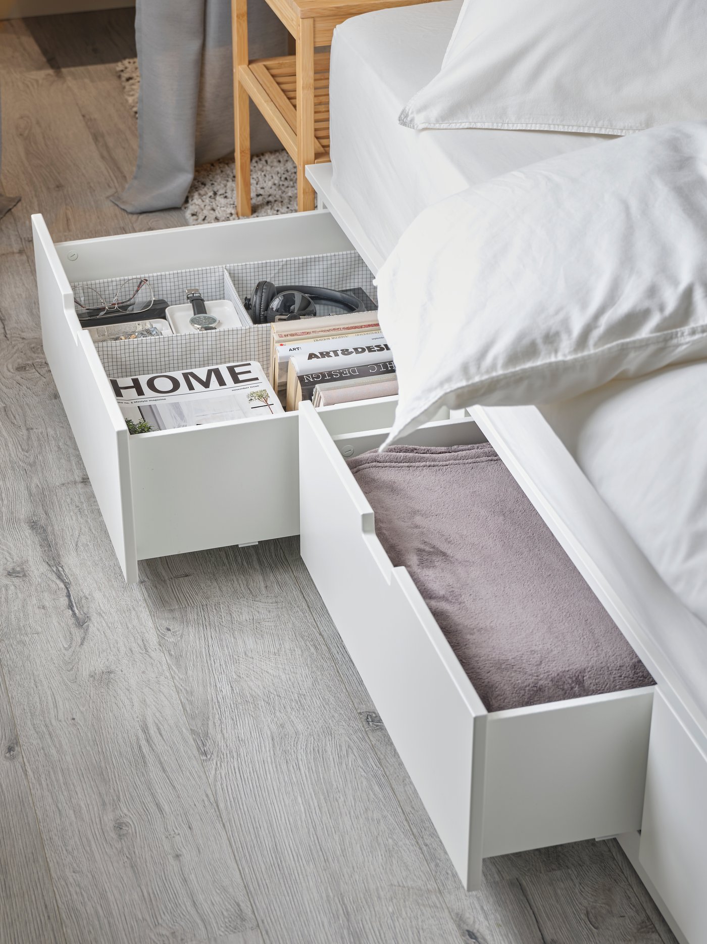 A white NORDLI bed frame with storage underneath. Two of the drawers are open and contain books, a bedspread and various items.