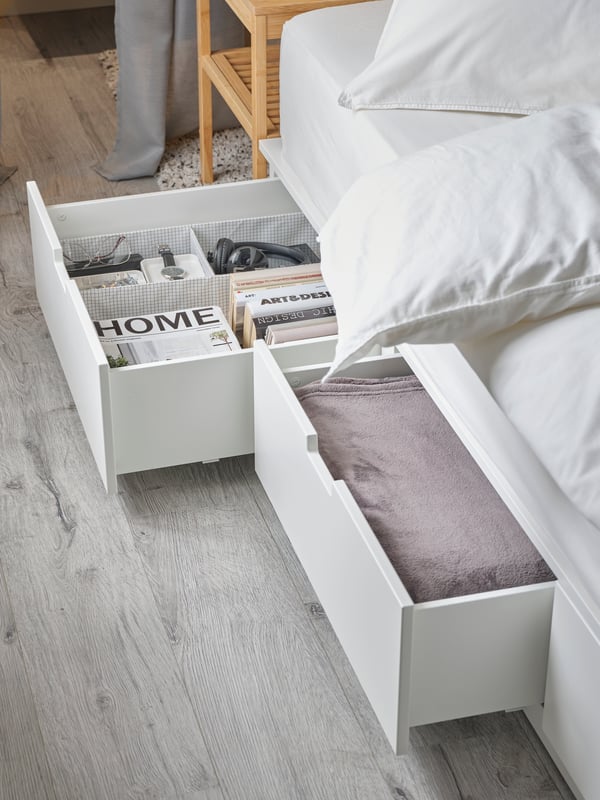 아래에 있는 화이트 NORDLI 노르들리 수납침대프레임에 두 개의 서랍이 열려 있고 책, 베드스프레드, 다양한 물건이 들어있는 모습.