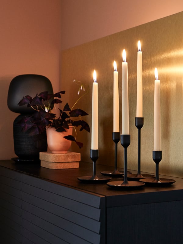 Lit candles in black candle holders, a potted plant and a speaker lamp, with a LYSEKIL brass-coloured wall panel behind.