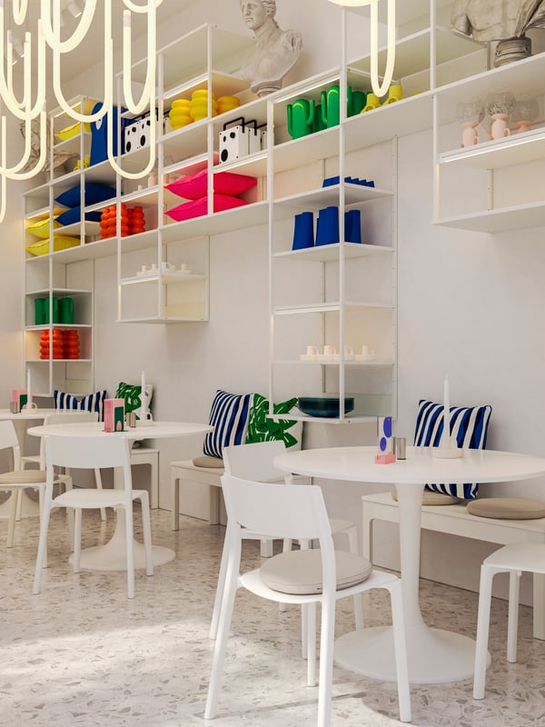 Un bar con tavoli e sedie bianchi e scaffali a giorno PLATSA montati a parete su cui sono esposti prodotti decorativi per la casa dai colori vivaci.