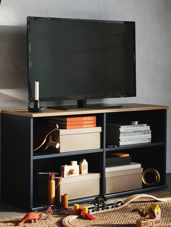 TV berwarna hitam berada di atas kabinet TV SKRUVBY berwarna hitam-biru dengan kotak, buku, mainan dan raket pingpong pada para-paranya.