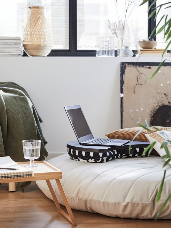 A black and white laptop support with a laptop on it, a natural pouffe, a tray, a glass of water, a notebook.