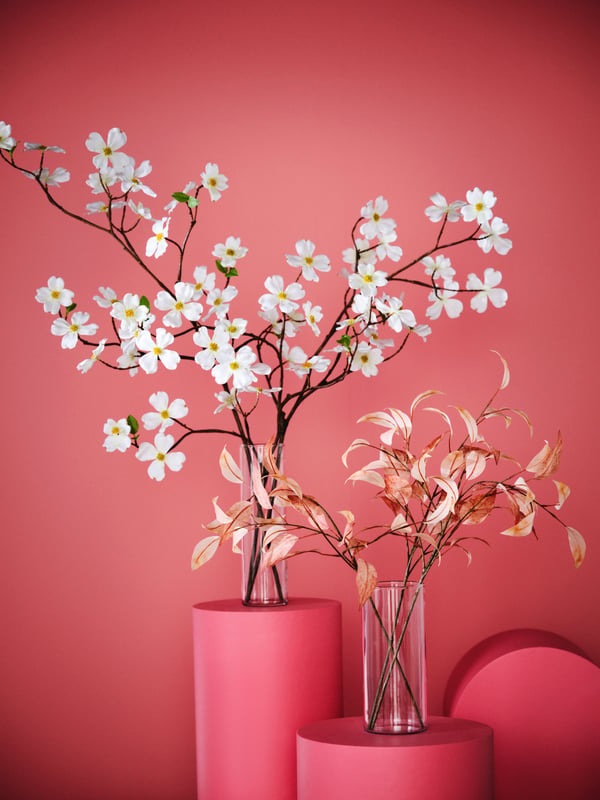 SMYCKA artificial cherry blossom with pink leaves sits in glass vases on a dark pink cylinder in front of a pink wall.