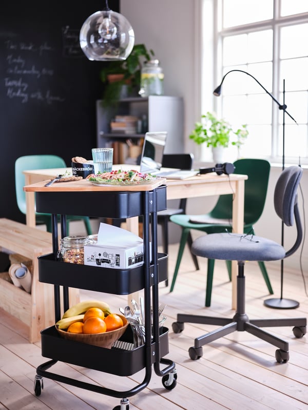 Carrello RÅSKOG nero con cibo e accessori per la tavola accanto a un tavolo INGO in pino. Al soffitto, lampada a sospensione JAKOBSBYN.