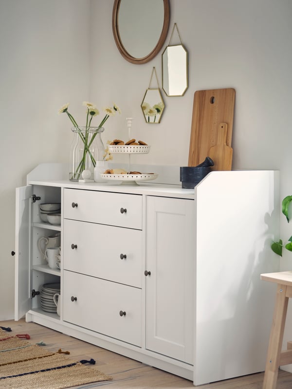 Flowers in a vase, a serving stand with cookies and chopping boards on a white sideboard. Wall-mounted mirrors above.
