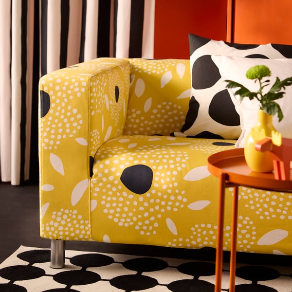 A white and yellow floral-patterned KLIPPAN cover for 2-seat sofa on a sofa sitting on a dotted black and white rug.