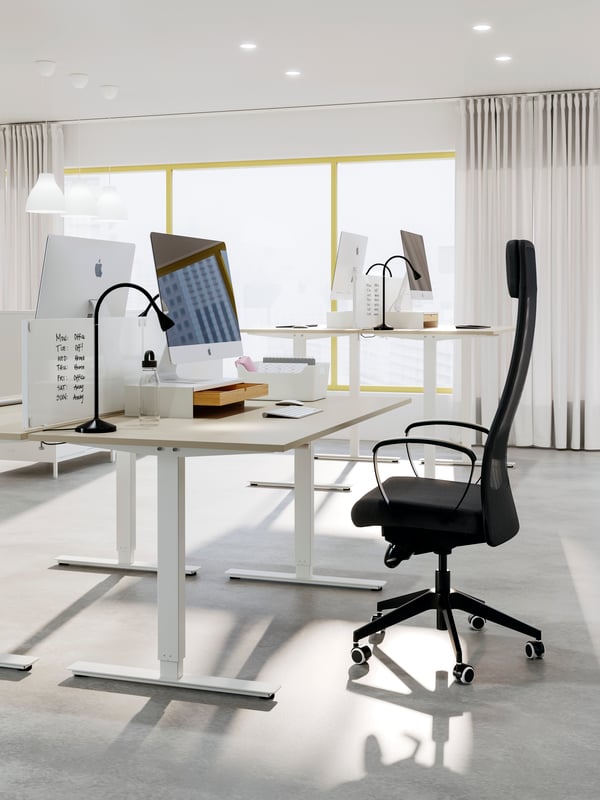 Scrivania regolabile in altezza TROTTEN beige/bianco con sedia grigio scuro e lampada da scrivania nera in un ufficio arioso.