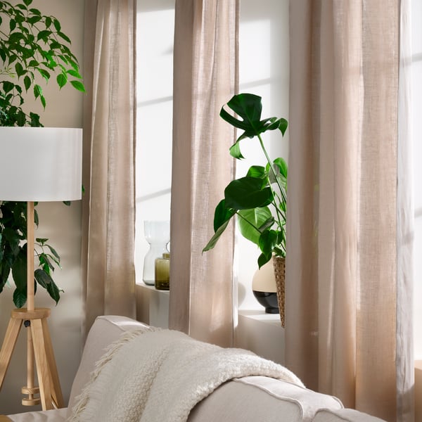 Dans une pièce ensoleillée, des plantes vertes et des vases sont disposés sur les appuis de fenêtres, encadrés par des rideaux DYTÅG beige foncé.