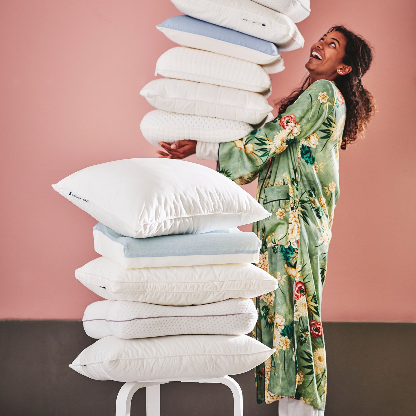 Personne portant une grande pile d’oreillers ergonomiques. Elle est derrière une pile plus petite, dont un oreiller SKOGSFRÄKEN sur le dessus.