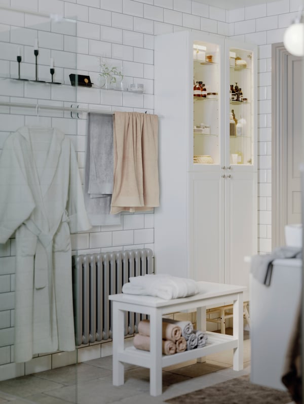 The corner of a tiled bathroom with towels and a bathrobe on two towel rails on the wall beside two GODMORGON high cabinets.