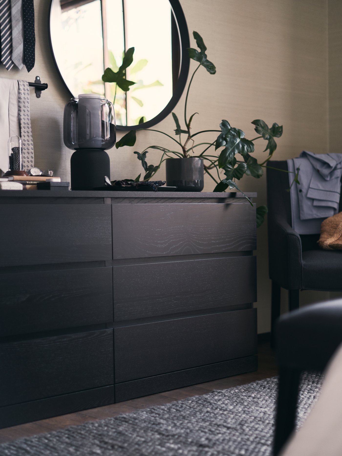 A black MALM chest of drawers with plants and a SYMFONISK WiFi speaker lamp on top. A mirror and tie rail on the wall behind.