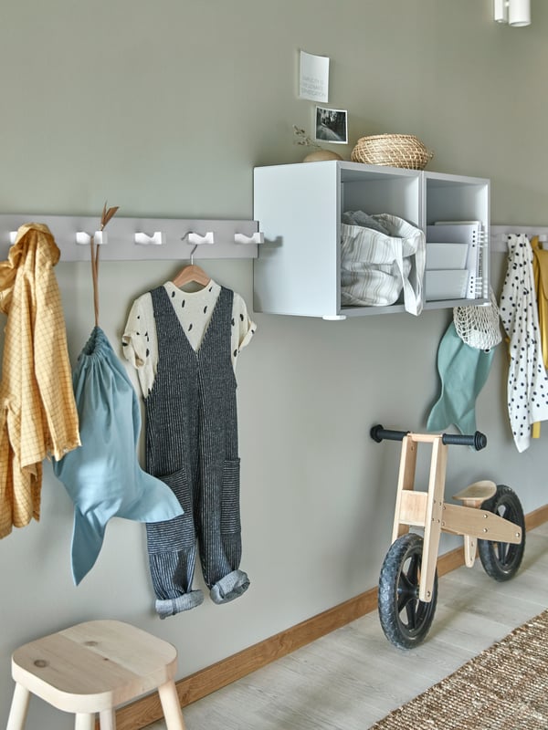 EKET shelving and KUBBIS hook racks on the wall holding children’s clothing, with a small bicycle and stool underneath.