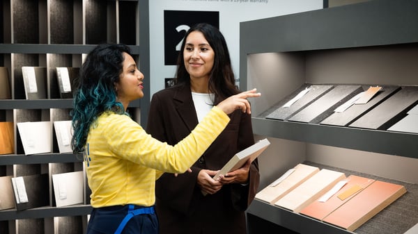 An IKEA co‑worker and a customer stand together in an IKEA store, reviewing and discussing kitchen material samples displayed on shelving during a planning consultation.