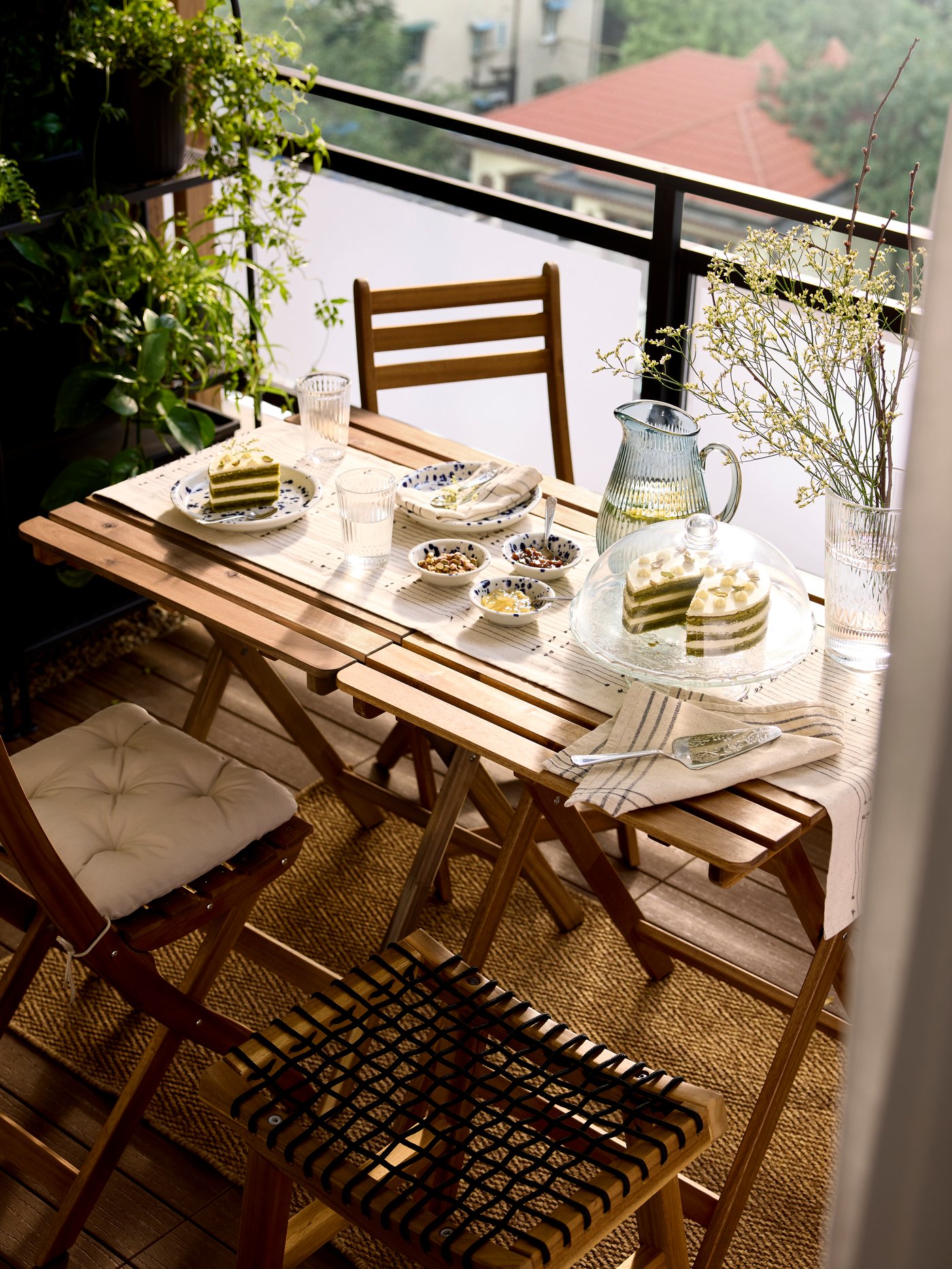A clear glass ARV BRÖLLOP serving stand with lid holds a matcha cake near plates on a small dining setup in a lush balcony.