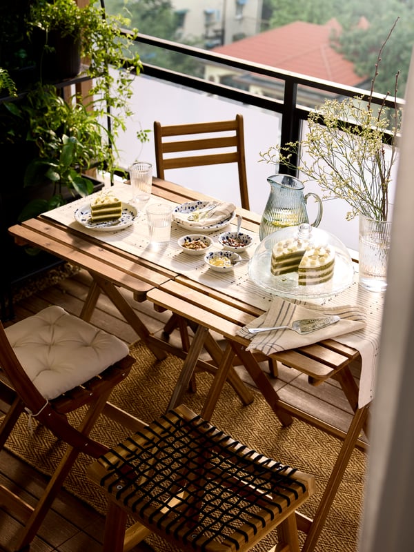 Ein Servierständer aus klarem Glas mit Deckel von ARV BRÖLLOP hält einen Matcha-Kuchen neben Tellern auf einem kleinen Esstisch auf einem üppig begrünten Balkon.