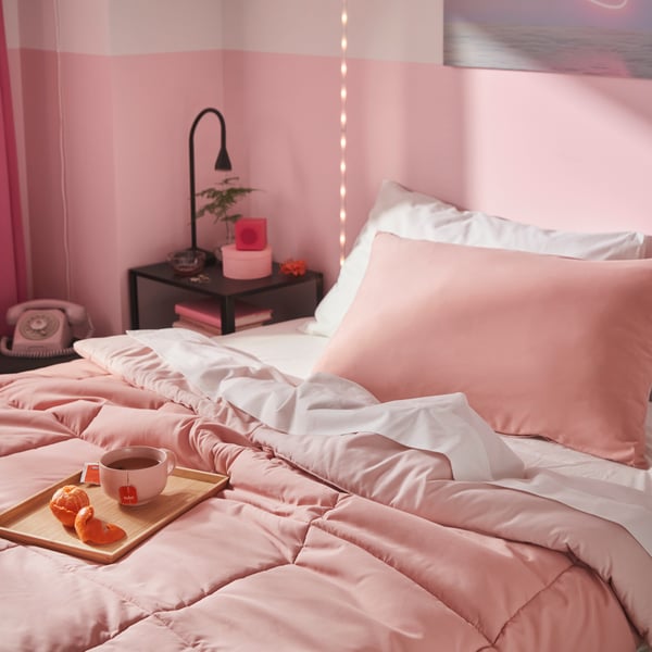 Una camera da letto decorata con un set di piumoni rosa HEDFRYLE drappeggiati su un letto, accompagnati da un vassoio con tè e frutta.