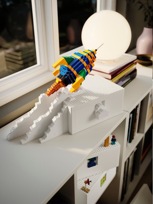 Un estante blanco y sobre él una serie de adornos entre los que destaca un cohete armado de LEGOS de colores.