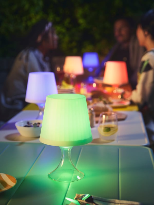 En soirée, espace extérieur où des personnes assises discutent, à la lueur de lampes LAMPAN en bleu, rouge et vert.