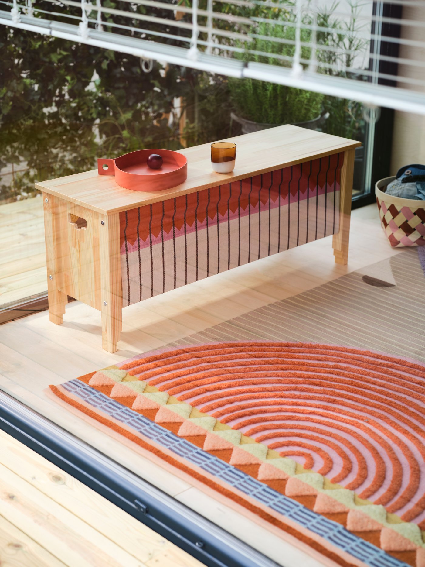 A multicolour OMMJÄNGE rug sitting on wooden flooring next to a bench with storage with textile weave on its long side.
