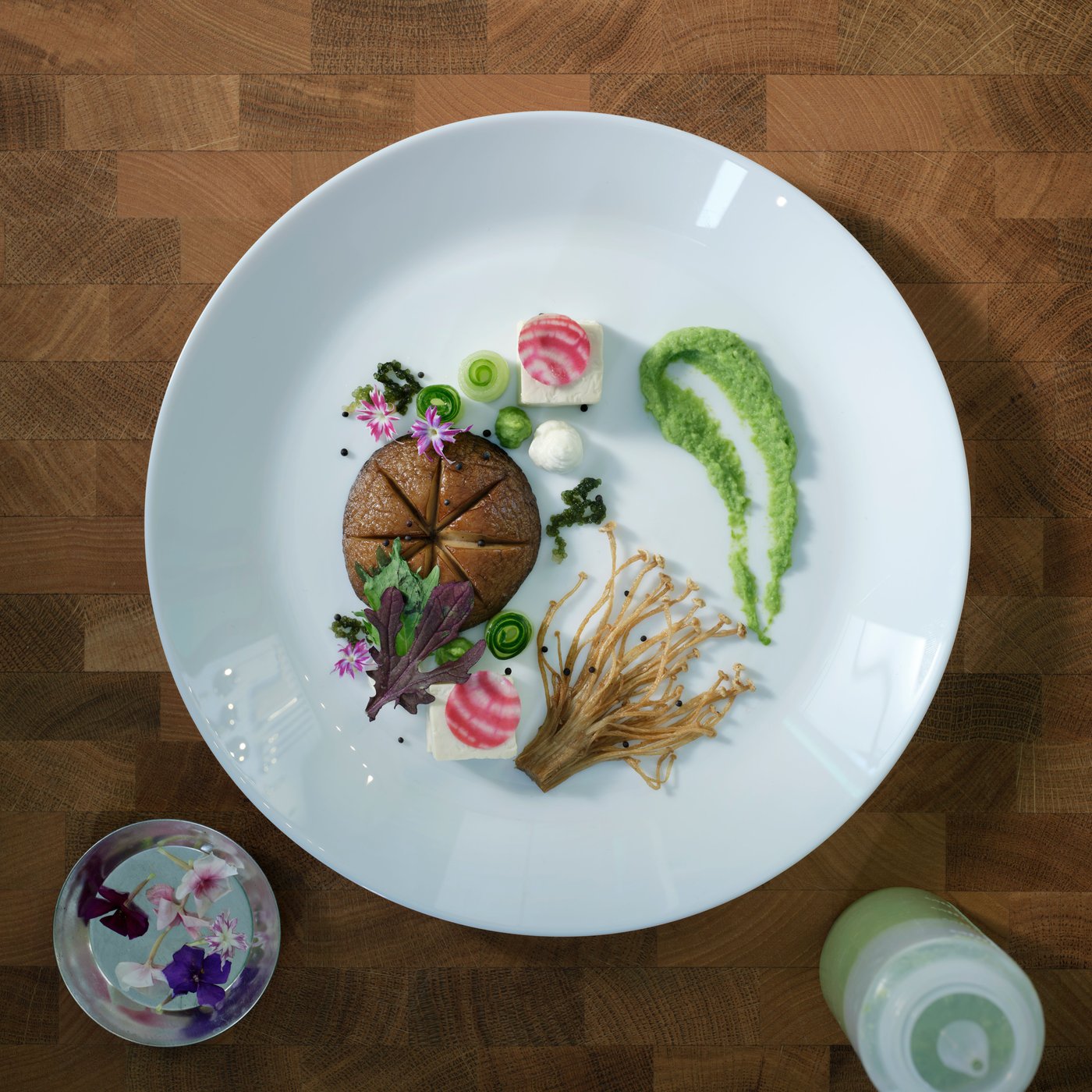 A white OFTAST plate is seen from above on a wooden surface. Food is beautifully arranged on it in a fine dining style.