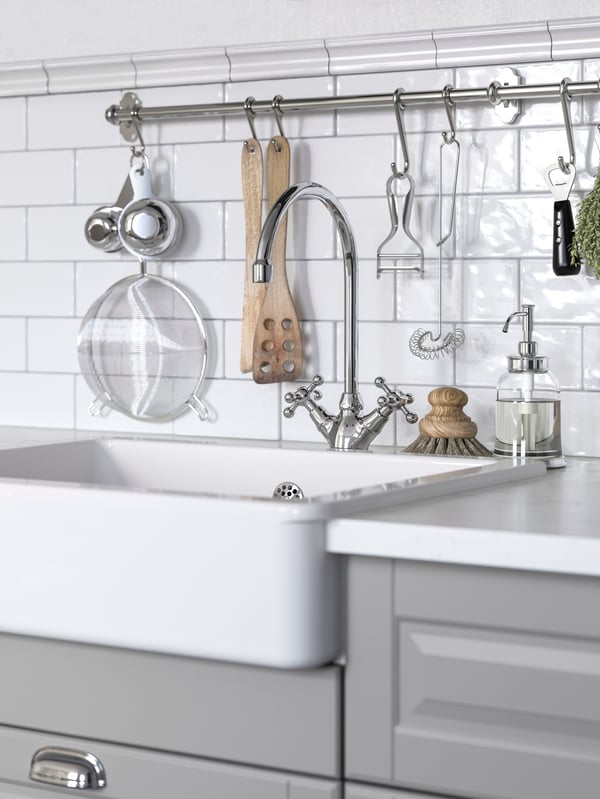 Vista de cerca de un lavaplatos de cerámica blanco sobre un mueble de cocina con cajones grises, empotrado a una pared con azulejos blancos. En la pared hay una barra metálicas de la que cuelgan diversos utensilios de cocina.