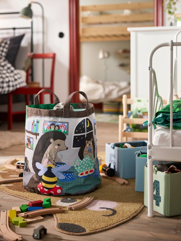 Close-up on SKOGSDUVA storage bag on the floor in front of a children’s bed, with toys scattered around it.
