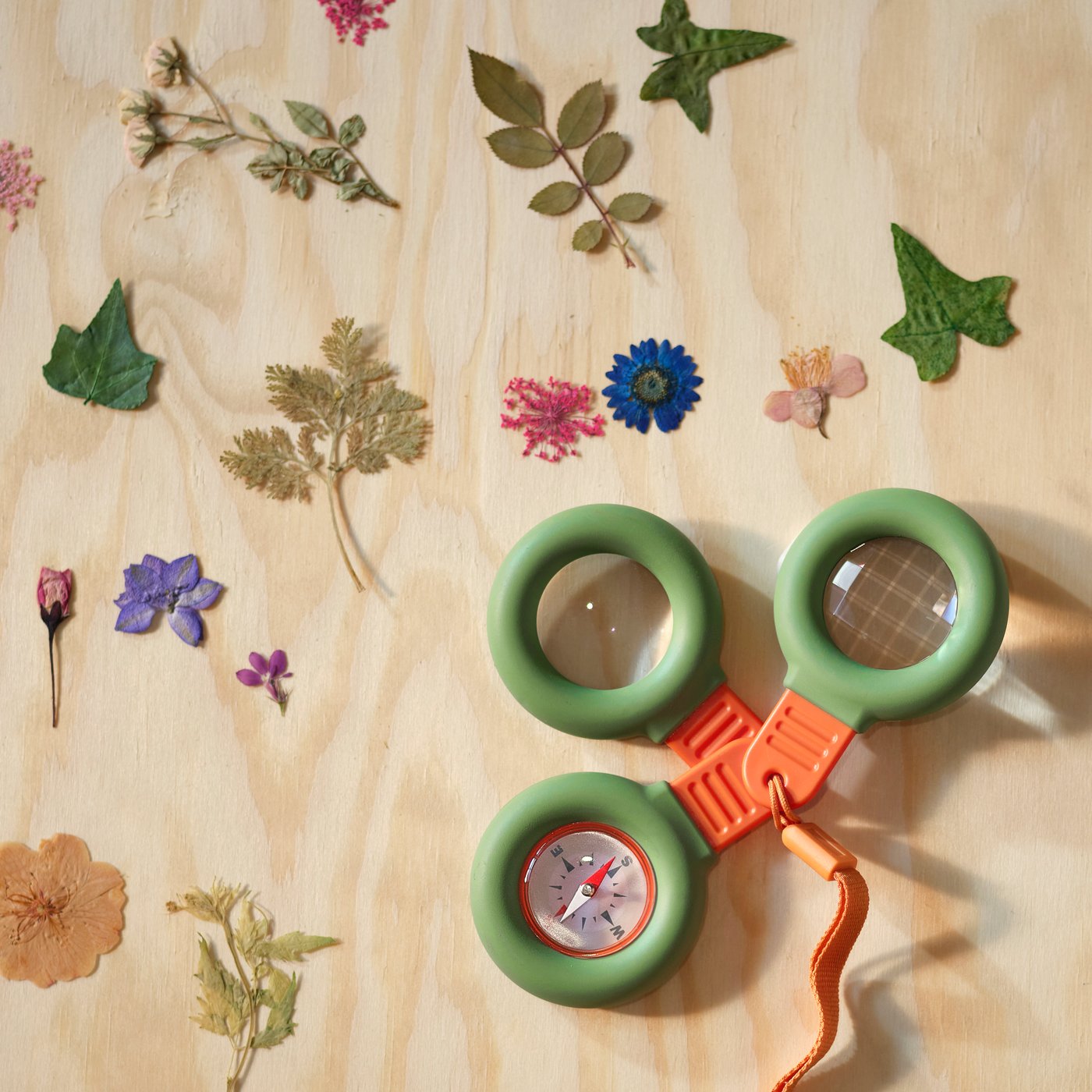 Un set de exploración MAJBAGGE con brújula, lupa y prisma colocado sobre una superficie de madera junto a flores secas.
