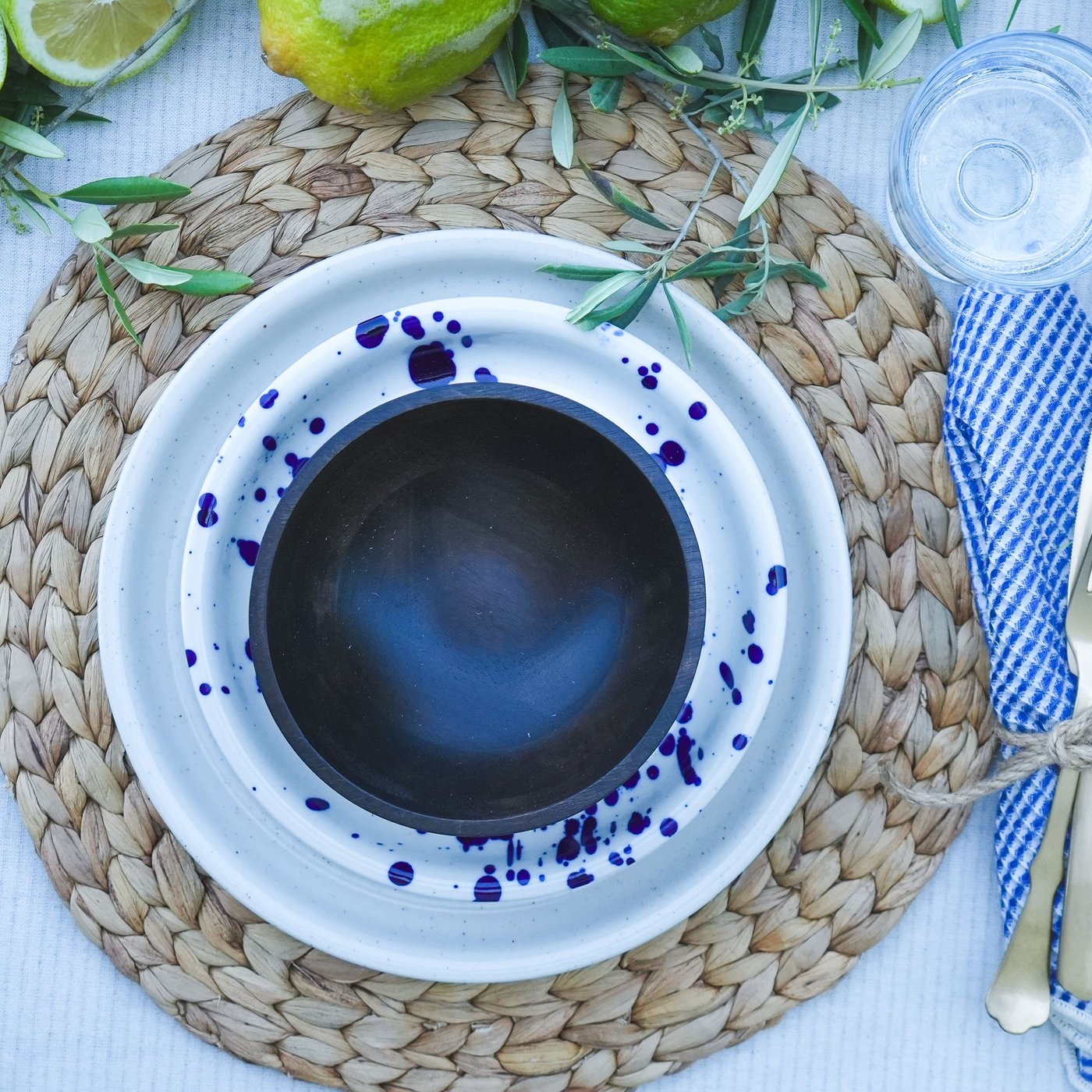Mediterraner Tisch gedeckt mit blau-weissem SILVERSIDA Geschirr und Besteck, umgeben von frischen Zitronen und Olivenzweigen auf einem geflochtenen Tischset.